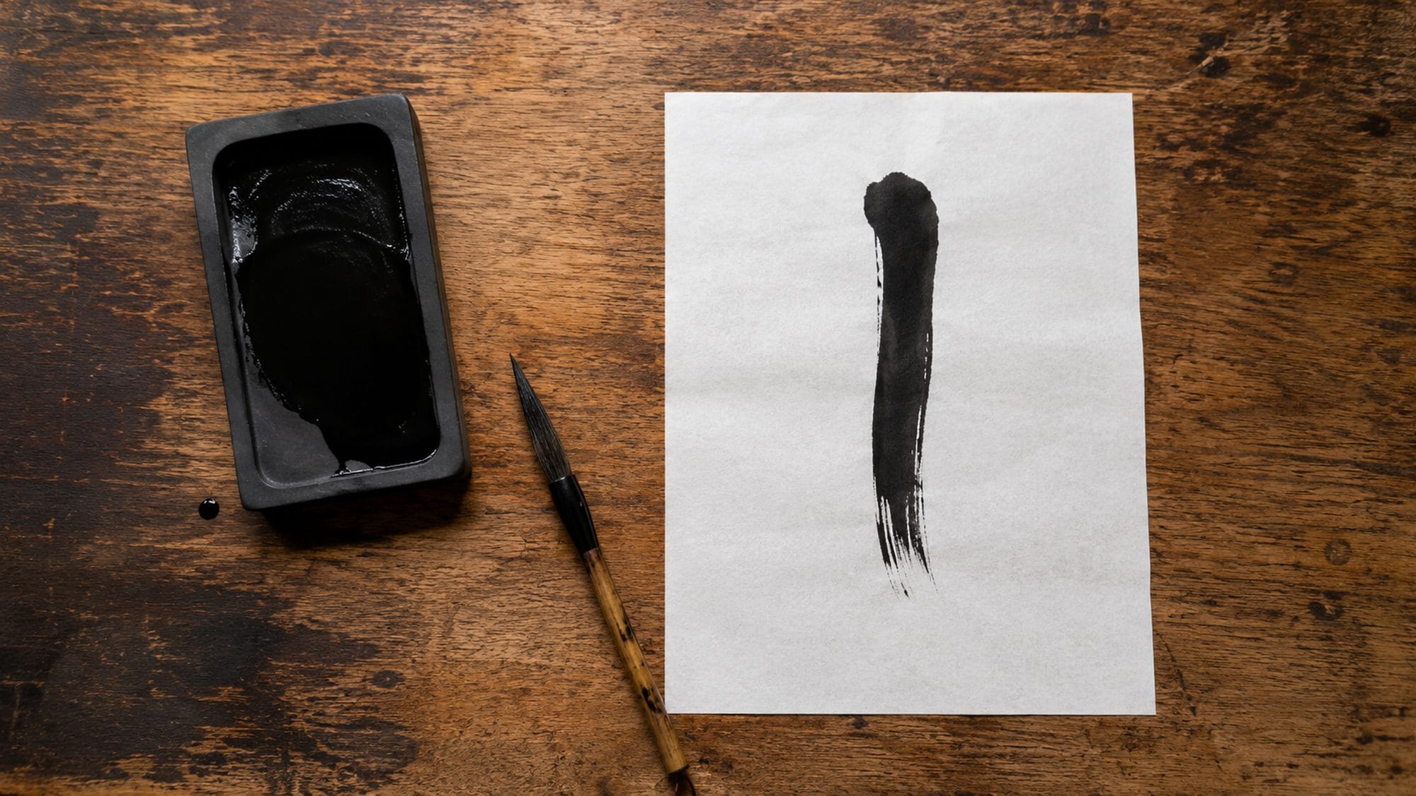 Overhead view of a Japanese calligraphy workspace — a suzuri inkstone with fresh ink, a bamboo brush, and a single brush stroke on white washi paper