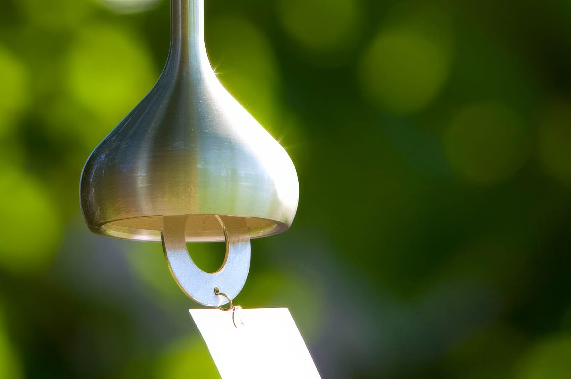 Nousaku tin wind bell hanging in a garden with soft green bokeh, showing the polished silver luster and teardrop shape of the finished casting