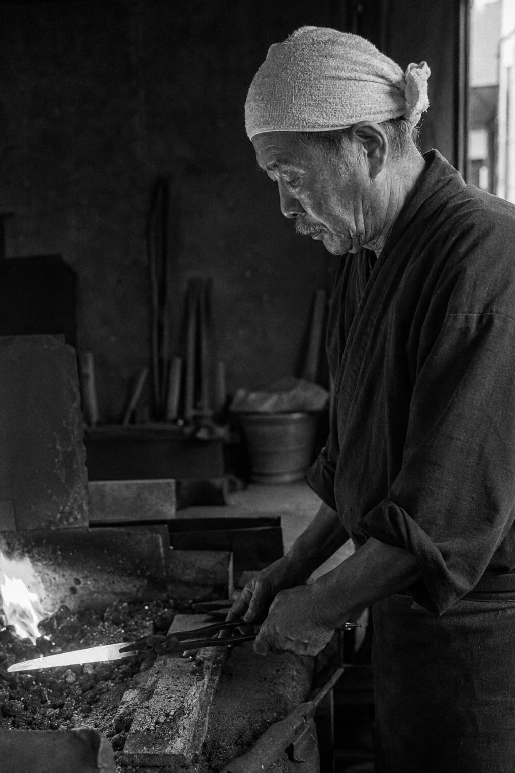 Japanese blacksmith in profile holding glowing steel with tongs over a charcoal forge