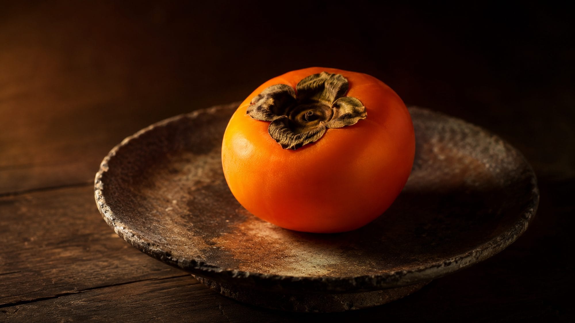 A ripe Japanese persimmon glowing deep orange on a dark aged ceramic plate with natural ash glaze