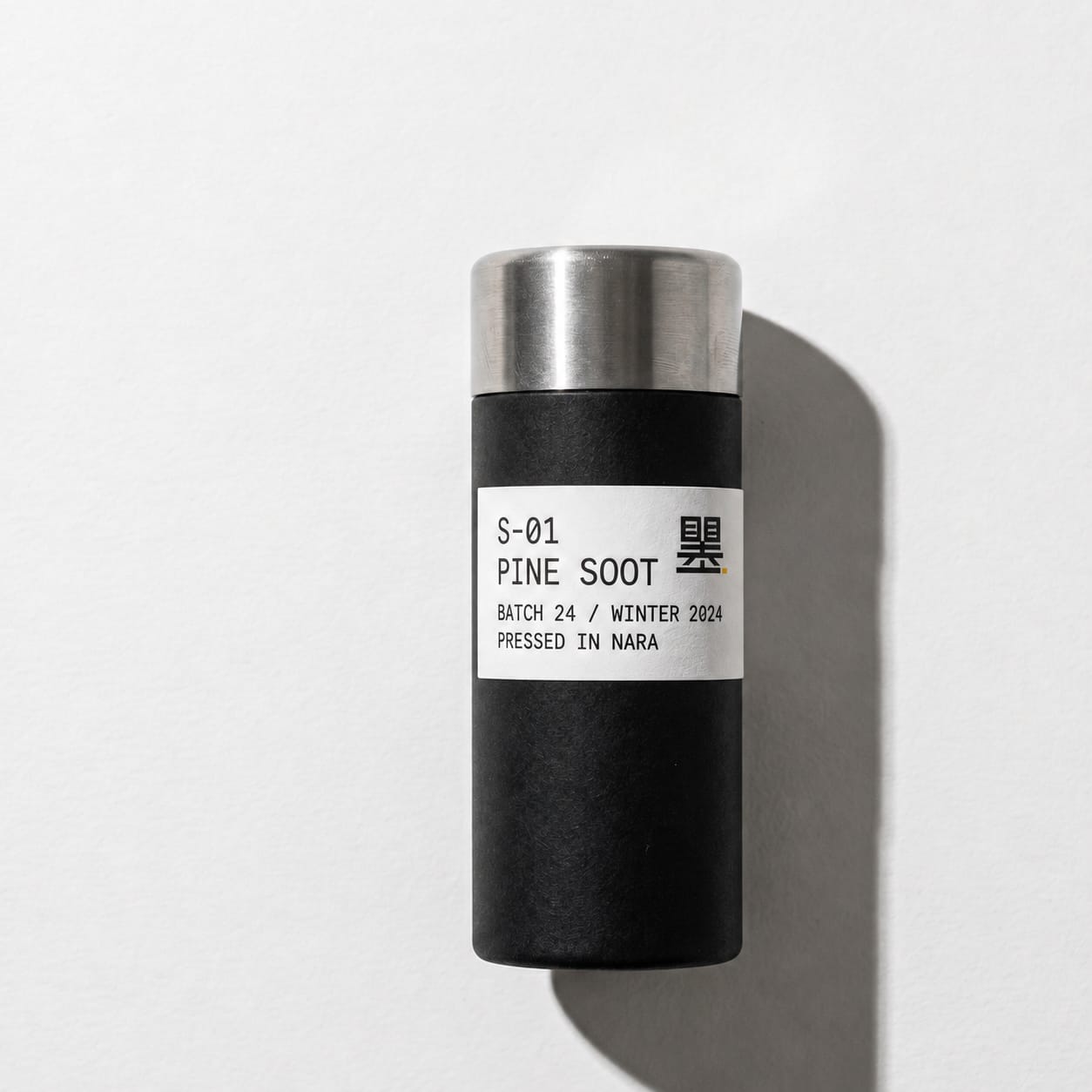 A matte black cylindrical packaging tube with brushed stainless-steel cap and wraparound monospace catalog label, photographed studio-flat on paper-white with a sharp directional shadow