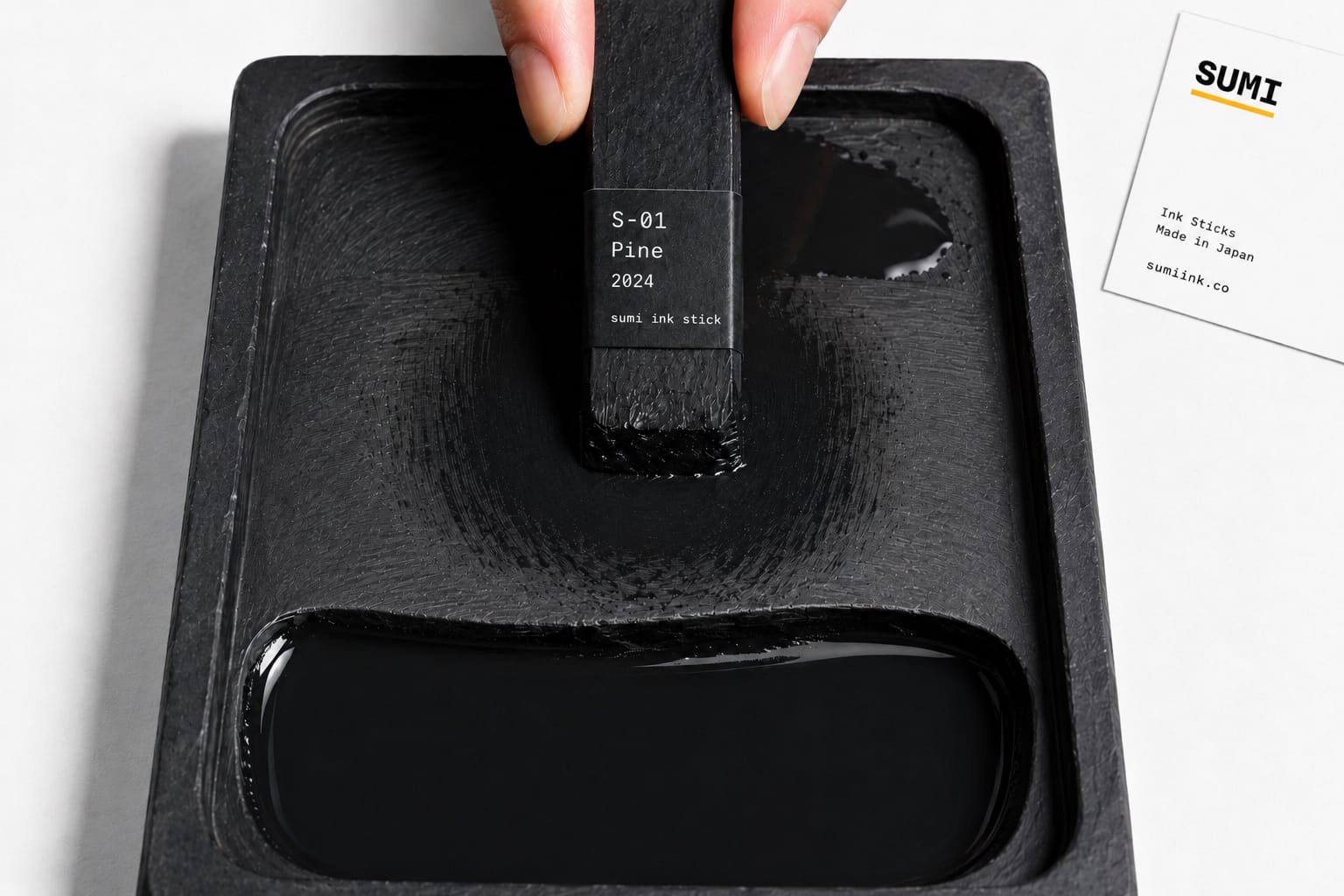 Close-up overhead view of a labeled sumi ink stick mid-grind on a slate-black suzuri stone, fresh ink pooled in the lower well, fingers gripping the stick from the top